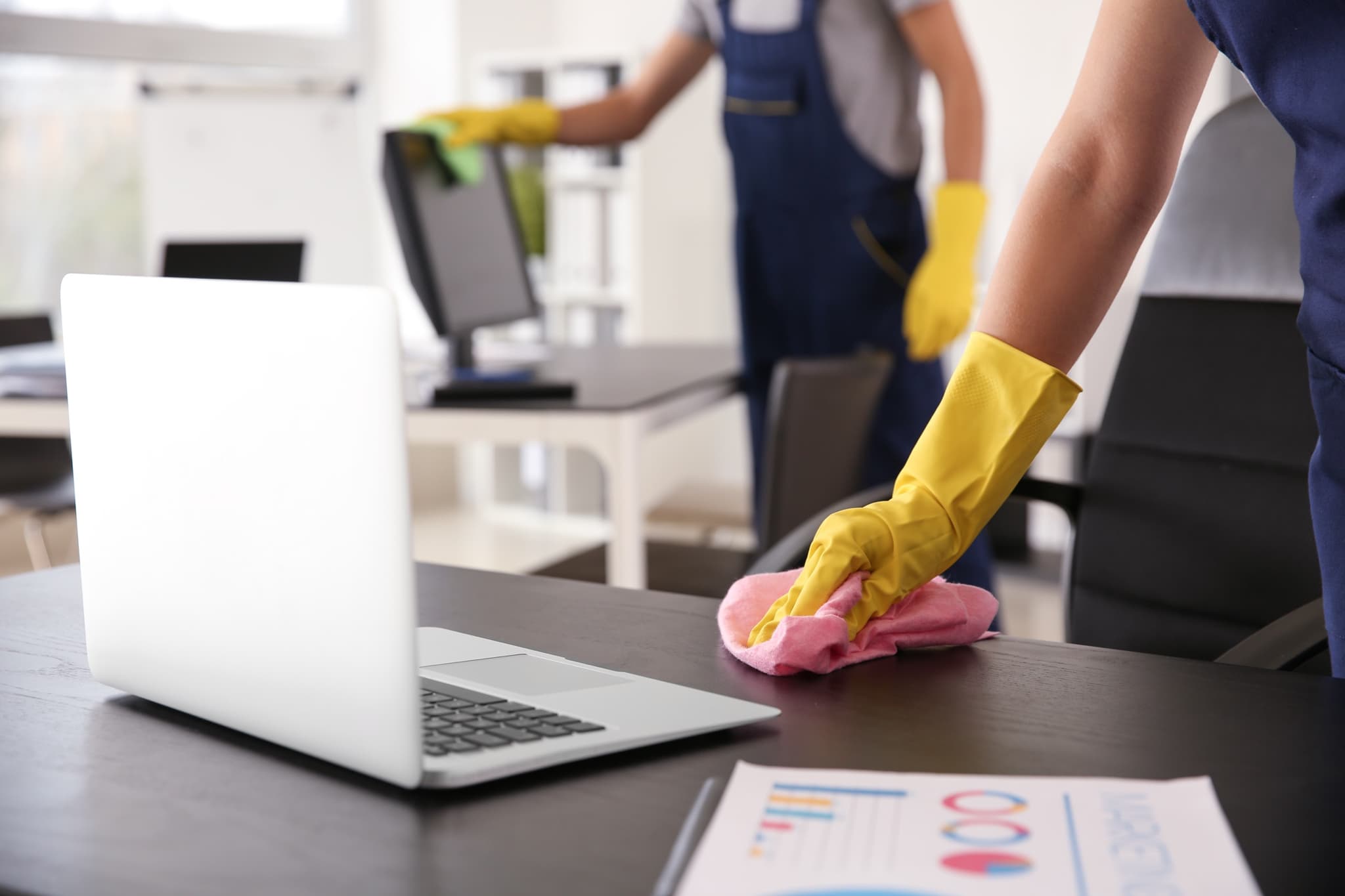 Úklidový tým v modrých pracovních overalech čistí kancelář - ruka ve žluté rukavici otírá stůl u laptopu, v pozadí kolega čistí monitor